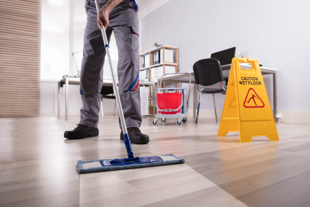 SCS Group cleaning service cleaning a wooden floor in an office setting. The man is seen using a mop to clean the floor, indicating that he is performing cleaning duties.